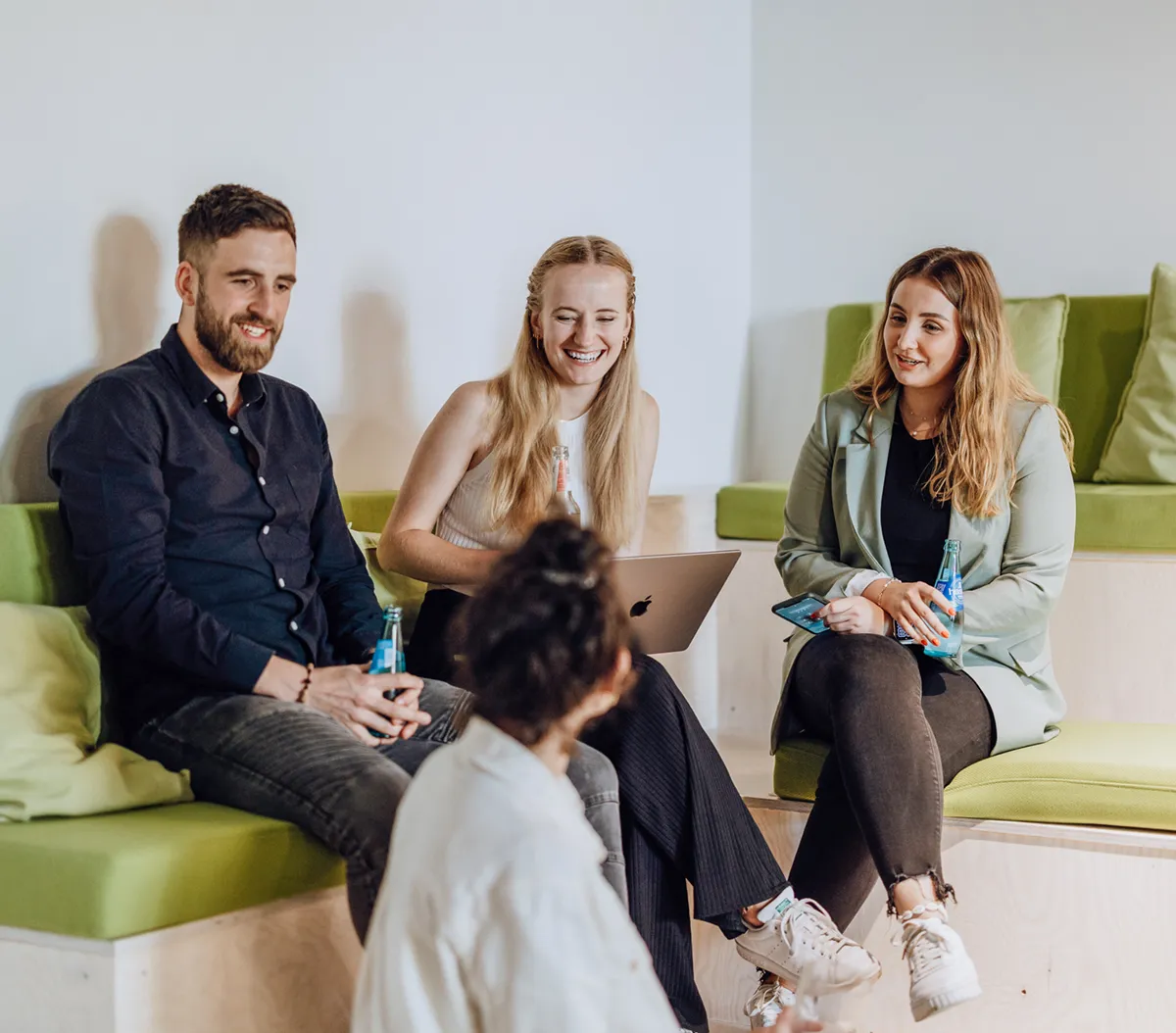 A group of four people sitting on green cushioned benches, engaged in a friendly conversation. One person is holding a laptop, while others have drinks in their hands. They are smiling and appear relaxed in a casual setting.